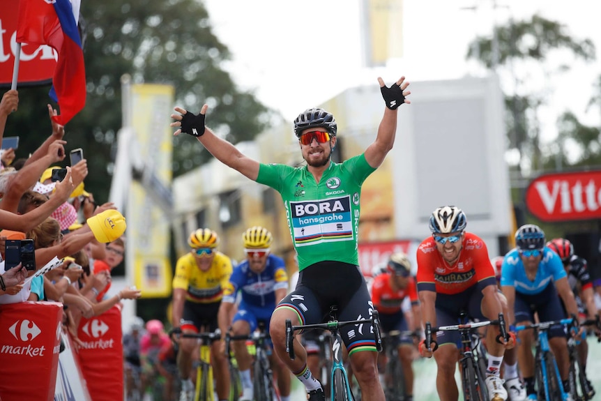 Peter Sagan holds his hands up wearing a green jersey as other riders cycle behind him