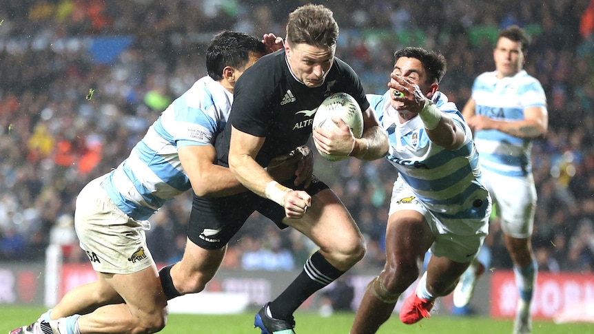 An All Blacks player scores a try as he is tackled by two Pumas players.