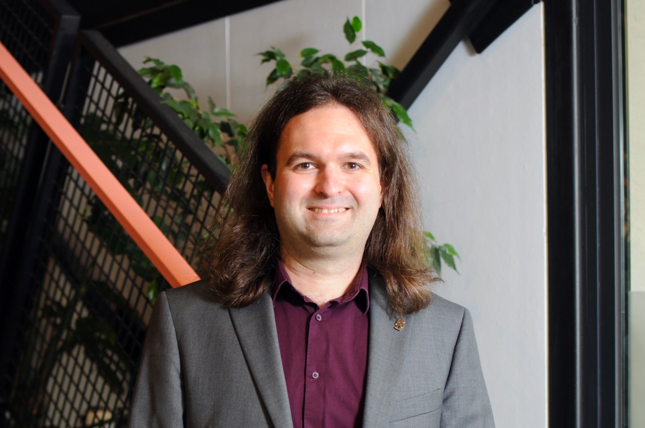 A man wearing a blazer smiles standing in front of some stairs.