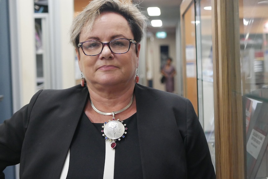 Woman with glasses and large pendant on a necklace looks at the camera.