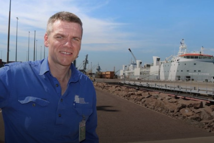 A man in a blue shirt stands in a boat harbour with a ship behind him