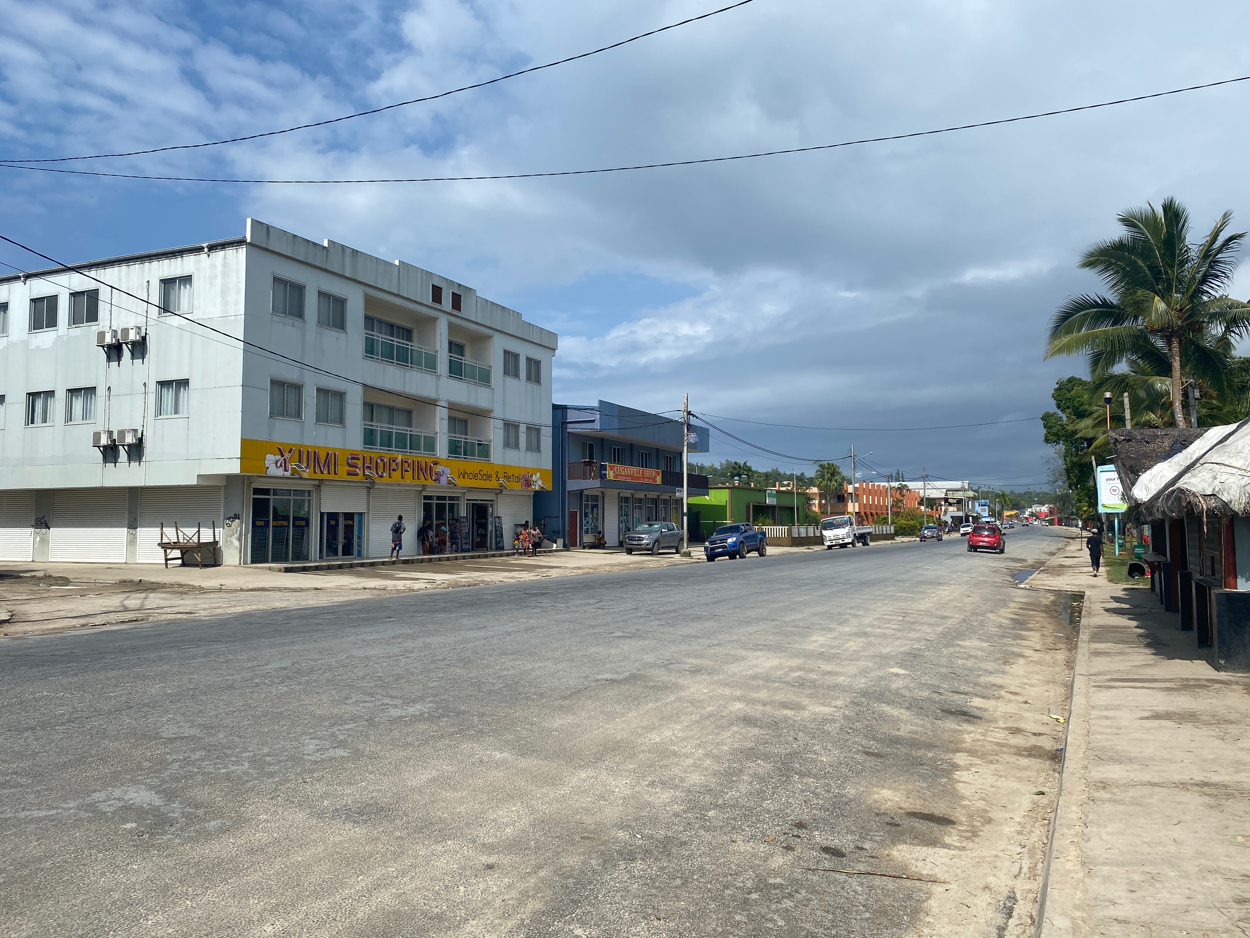 Uma rua larga, empoeirada e asfaltada, com alguns carros estacionados no meio-fio e um carro se afastando ao longe.