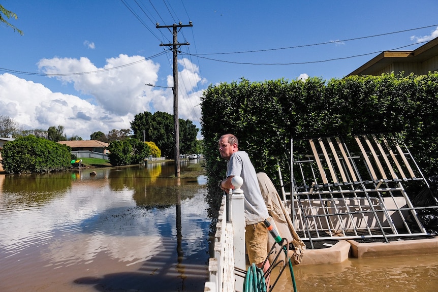 A man leans on a fence looking at brown floodwaters filling his street