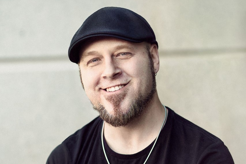 A man in black t-shirt with facial hair wears black flat cap hat, silver necklace and poses in front of concrete wall outdoors.