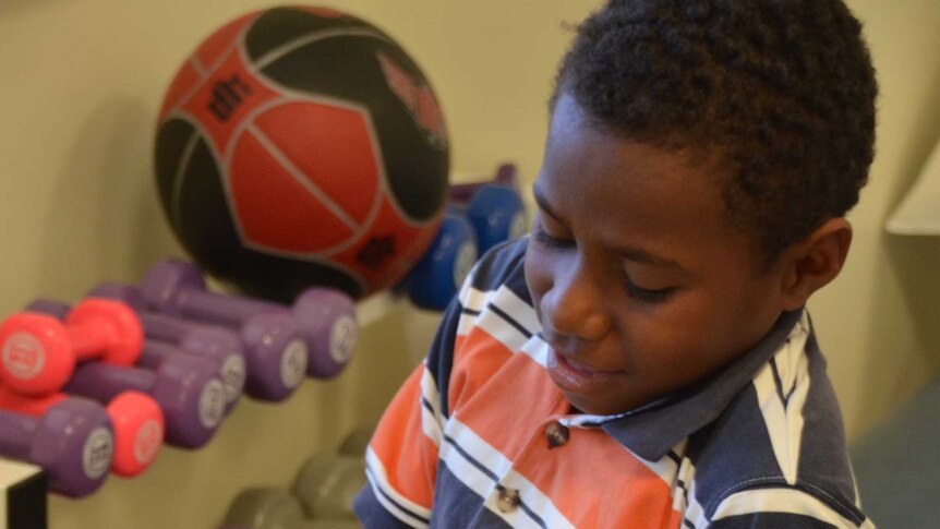 Boy from Papua New Guinea smiles as he tries a weight for exercise on his finger, weights and balls behind him.