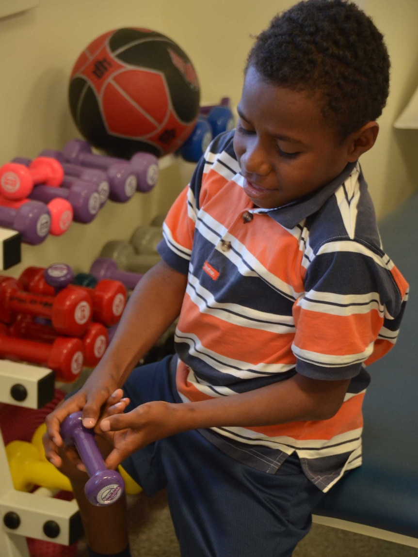 Boy from Papua New Guinea smiles as he tries a weight for exercise on his finger, weights and balls behind him.