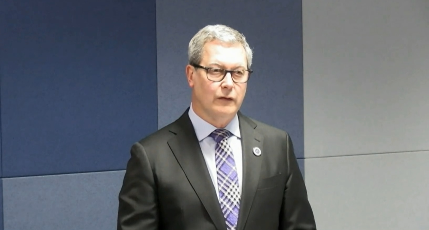 Mr Lancaster in a black suit and purple tie with lines crisscrossing on it standing and asking questions.