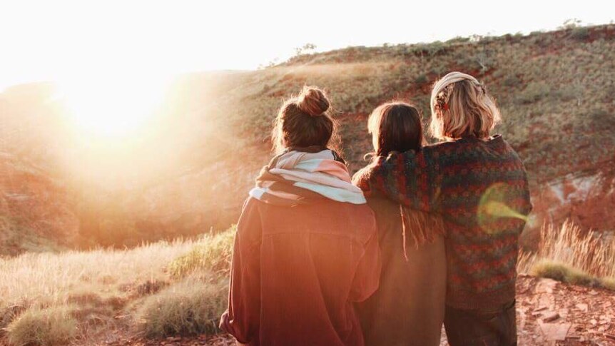 Three young people stand together watching the sunset over a hill