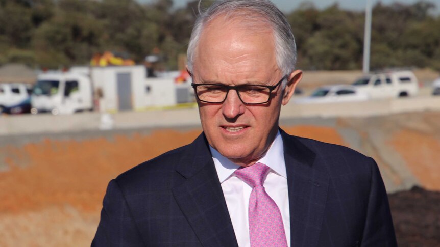Prime Minister Malcolm Turnbull looks downward standing outdoors wearing glasses and a suit with vehicles in the background.