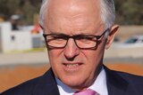 Prime Minister Malcolm Turnbull looks downward standing outdoors wearing glasses and a suit with vehicles in the background.
