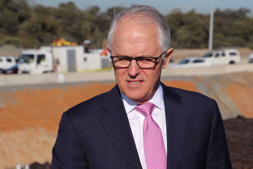 Prime Minister Malcolm Turnbull looks downward standing outdoors wearing glasses and a suit with vehicles in the background.