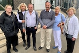 Two men in their fifties and sixties stand with their wives, and aunt and daughter outside court