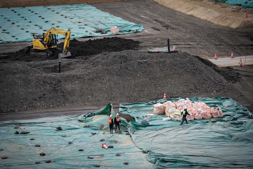 A yellow digger sits upon a large mound of black dirt with two workers in hard hats in the foreground