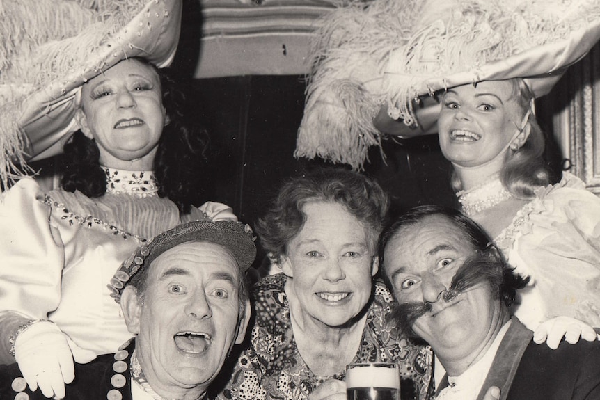 A black and white photo of two women wearing giant, fluffy hats, who are standing behind three other people in a bar