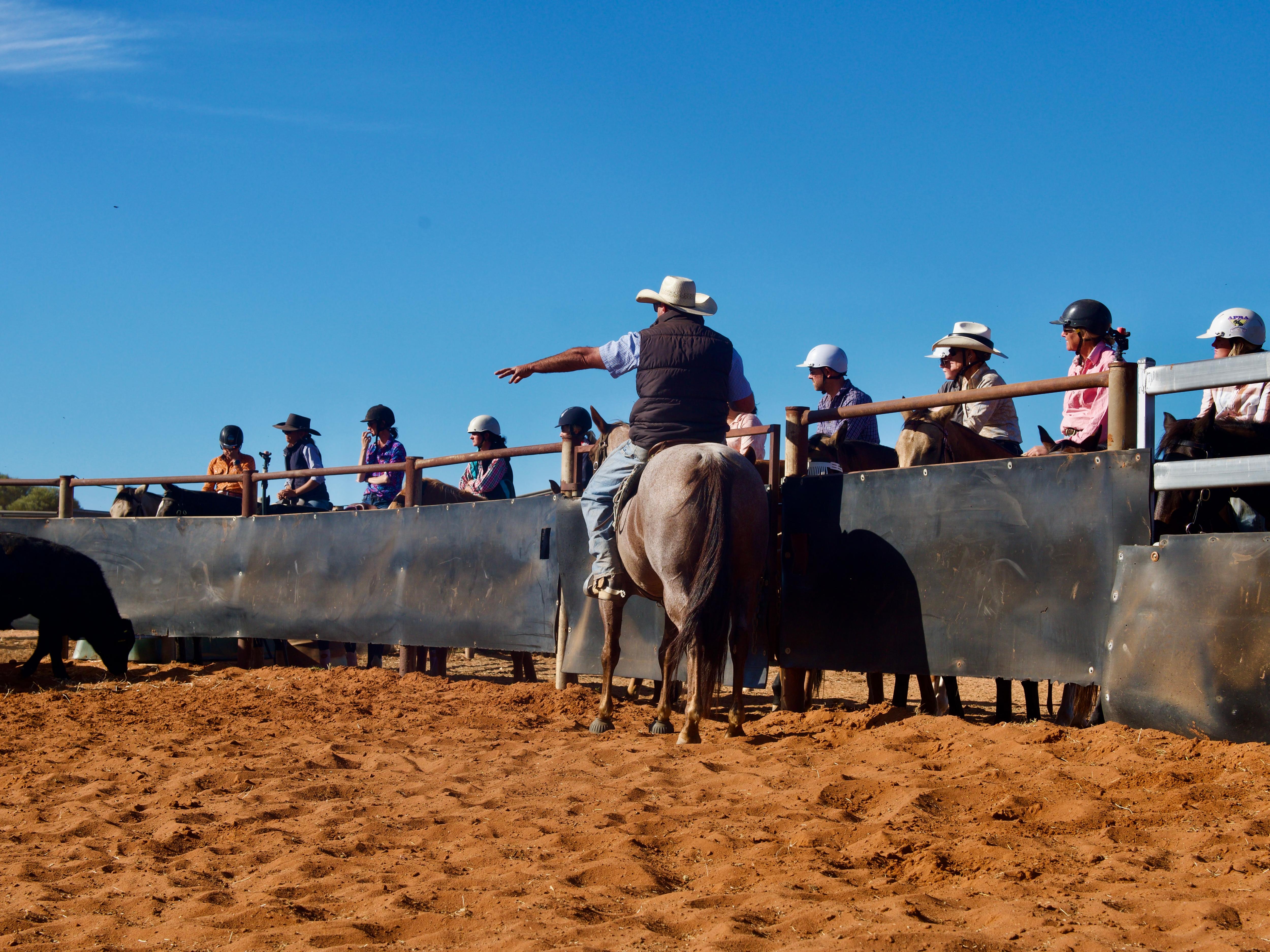 a man with a hat on sitting on a horse in a yard with red dirt pointing with other horse riders in the background 