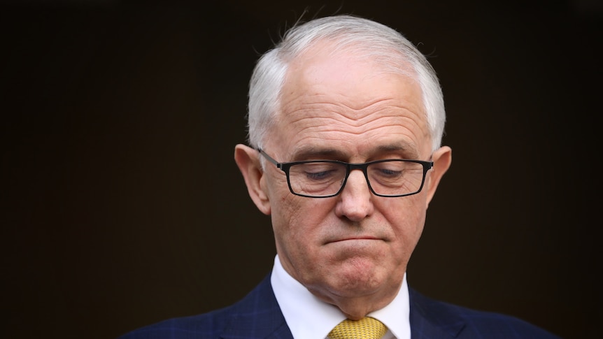 A bespectacled man in a blue suit with a yellow tie and grey hair looks down with clenched lips