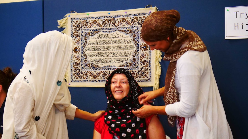 Two women wearing hijabs wrap a headscarf for another who is sitting between them.