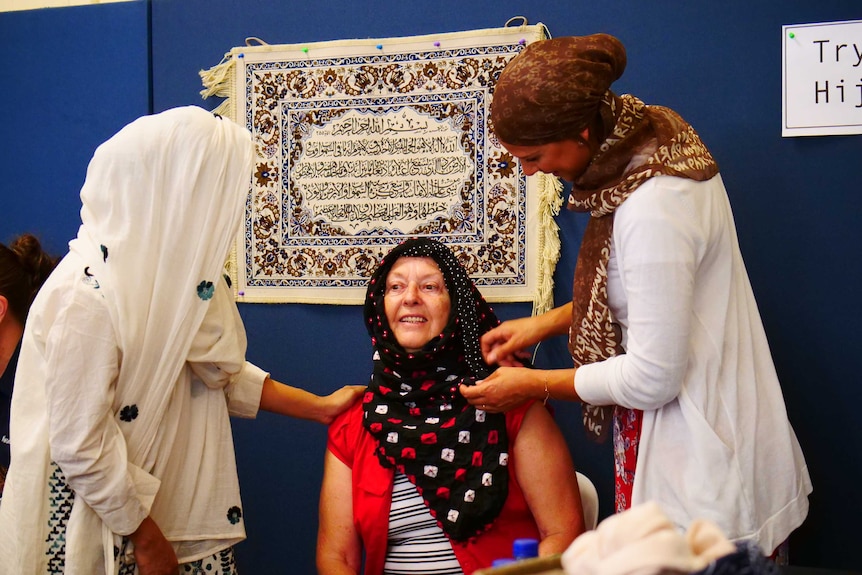 Two women wearing hijabs wrap a headscarf for another who is sitting between them.