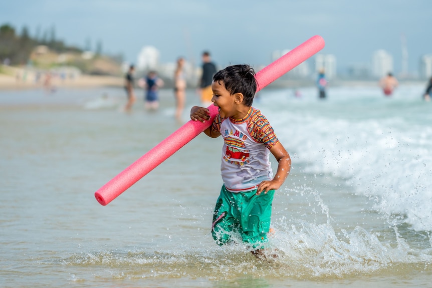 A four year old boy runs with a swimming noodle in the shallow water at the beach.