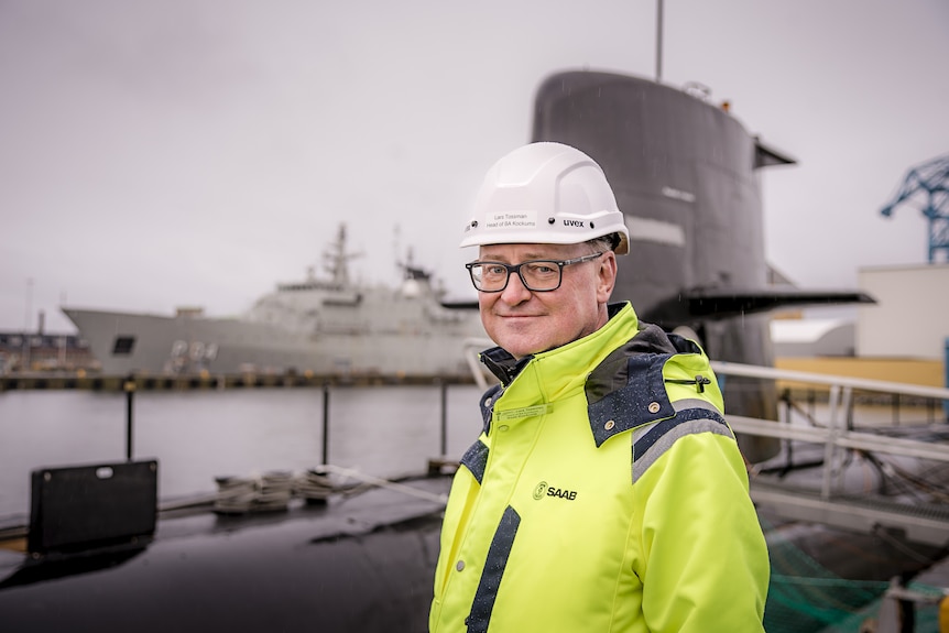 A man wearing a yellow jacket and white helmet stands next to a submarine.