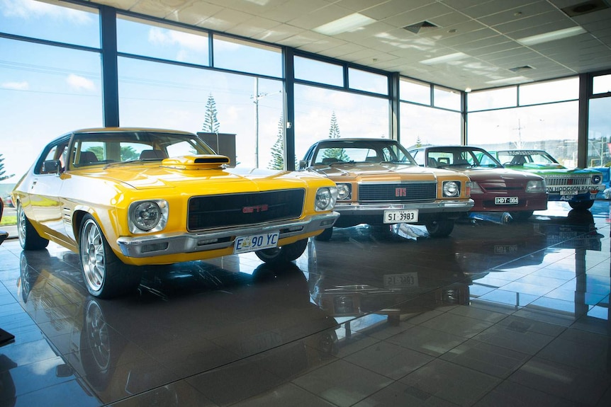 A line of four historic Holdens inside showroom. The closest is a HQ Monaro