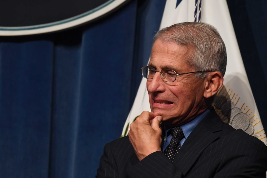 A bespectacled man with short grey hair pinches his chin and grimaces while standing in front of a flag and blue drapes.