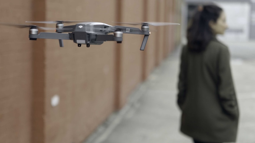 A drone flies just behind a woman as she walks down an alleyway.