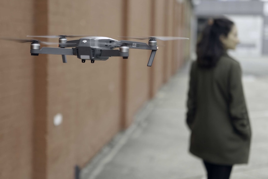 A drone flies just behind a woman as she walks down an alleyway.