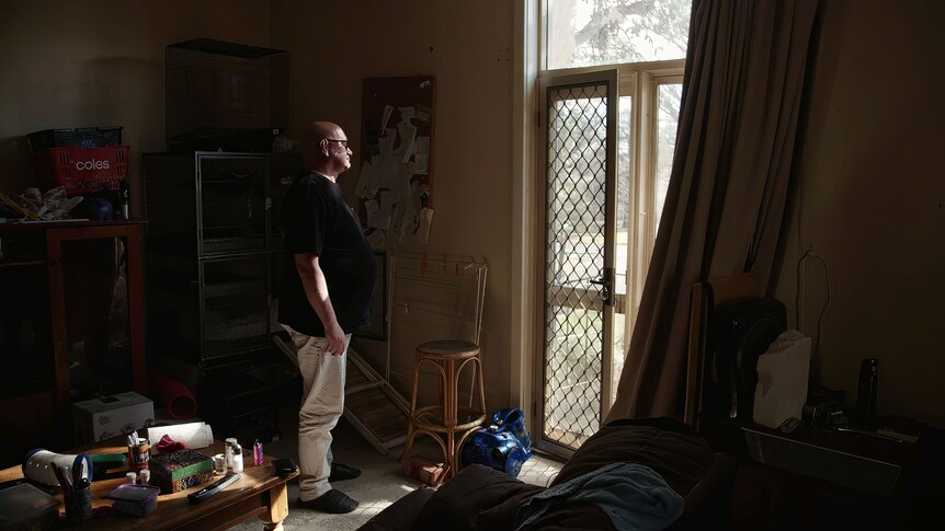 A man stands in a dark room looking out of a window 