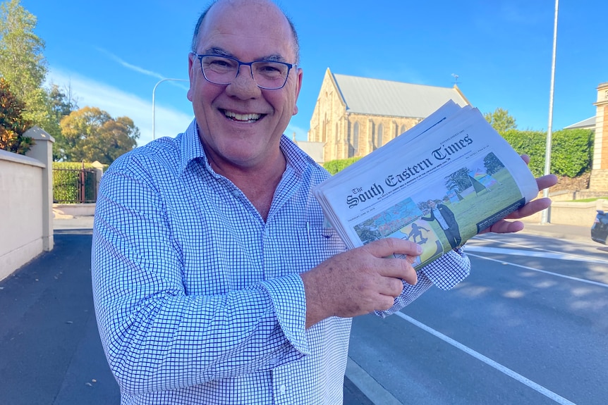 A man in a button up shirt smiles at the camera holding a newspaper