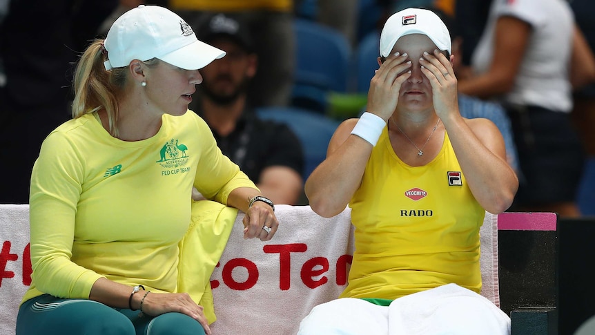 A captain sits on a bench talking to a female tennis player whose hands are covering her eyes.