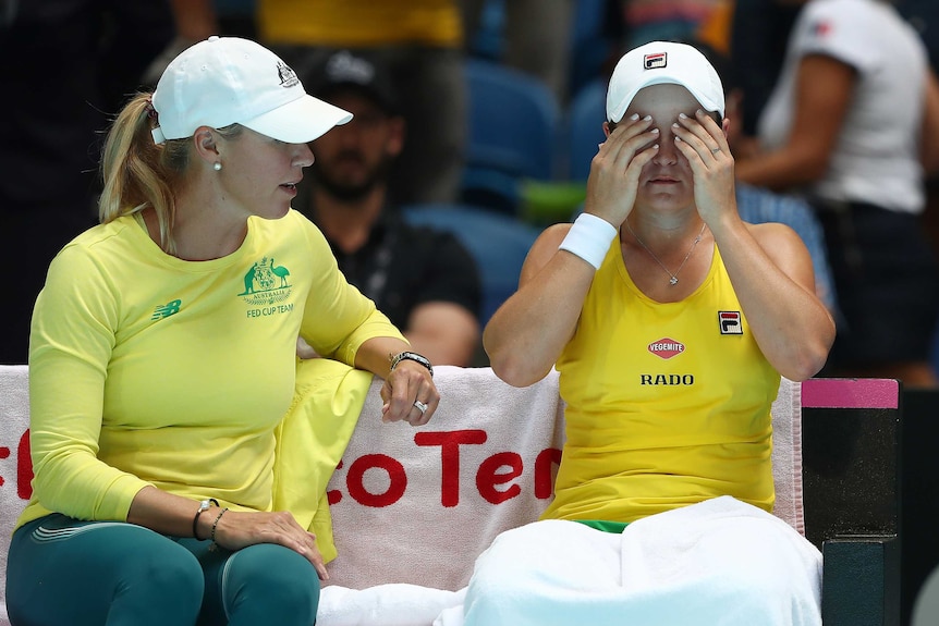 A captain sits on a bench talking to a female tennis player whose hands are covering her eyes.