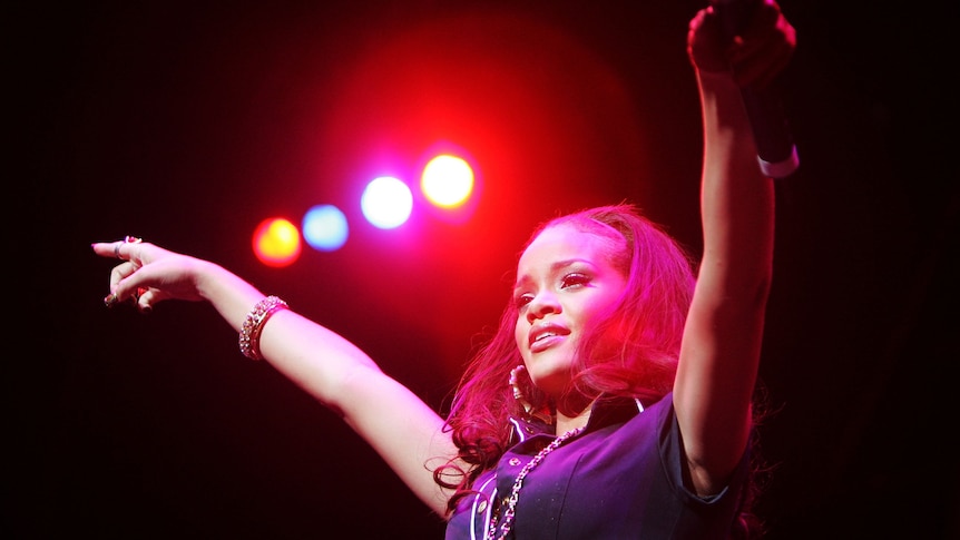 Rihanna stands on stage with both hands held up in the air. She holds a microphone in one hand.