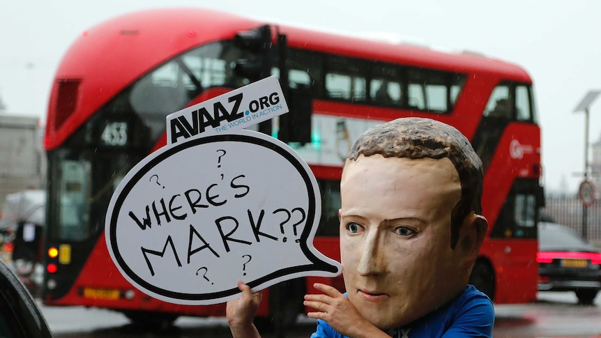 A protestor wears a Mark Zuckerberg mask holding a sign reading 'where's mark' in front of a London red double-decker bus.