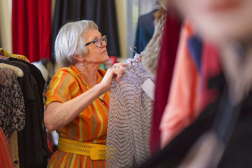 A woman in an orange and yellow striped dress and glasses hangs a frilly top from the rack.