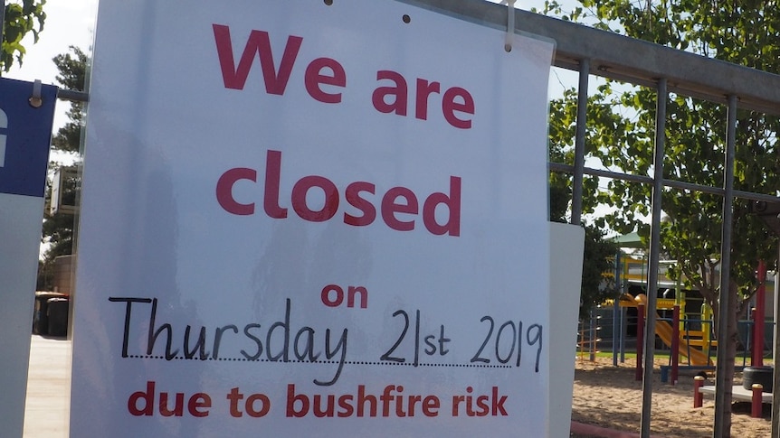 A sign secured to a metal school gate with zip ties reads 'We are closed on Thursday due to bushfire risk'.