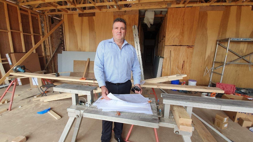 A man in a business suit stands in front of building plans, inside a house being built, with wooden frames visible.