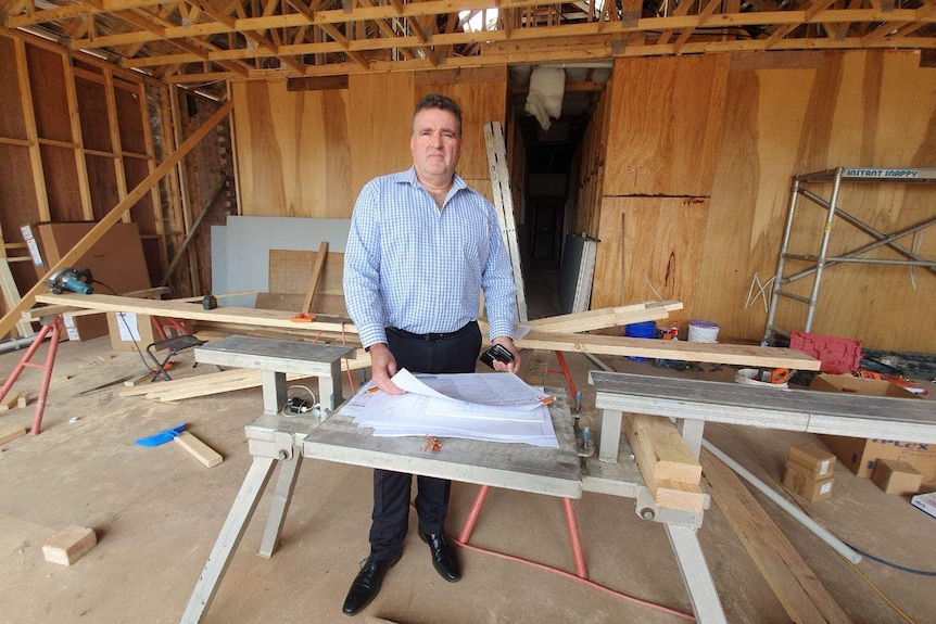 A man in a business suit stands in front of building plans, inside a house being built, with wooden frames visible.