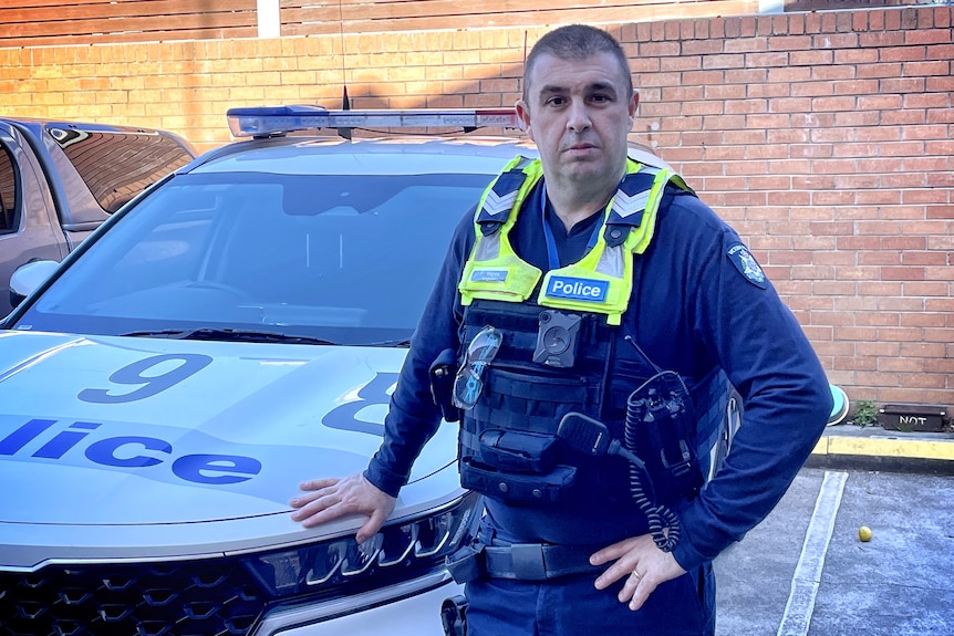 A police officer in uniform standing with one hand on the bonnet of his police car. 
