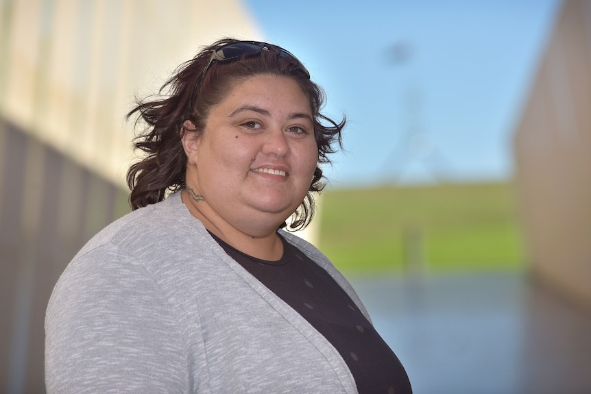 A woman with brown hair and a grey cardigan is smiling with Parliament House in the background