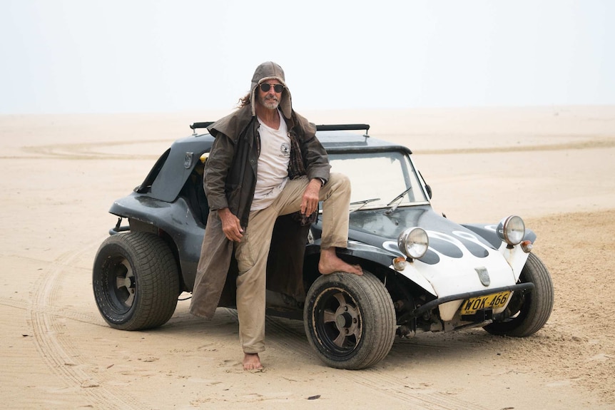 A beach buggy drives on sandy dunes between the ocean and a lake.