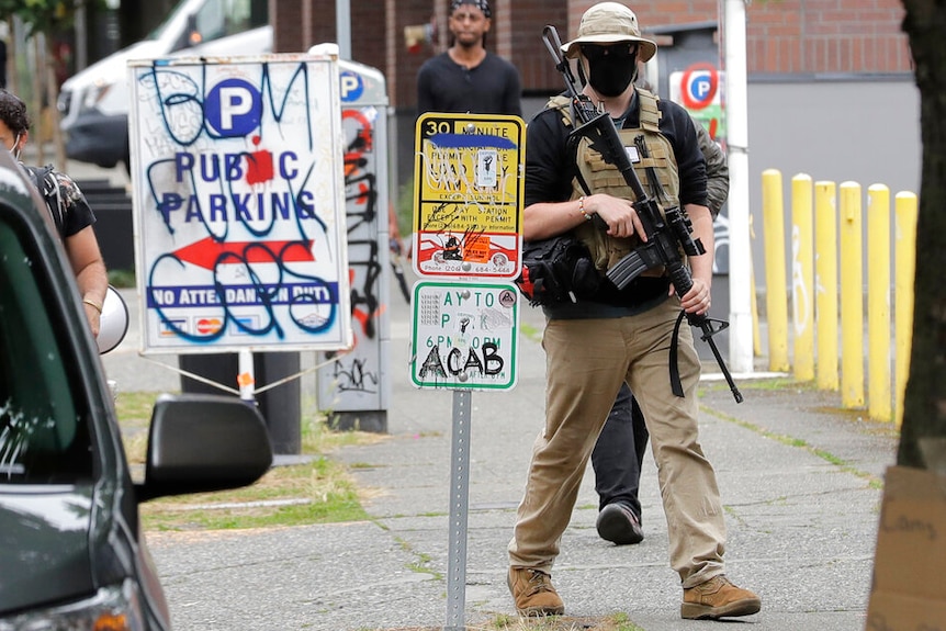 A man carrying an assault rifle while wearing a bulletproof vest, sunglasses, facemask and a hat walks along a Seattle street.