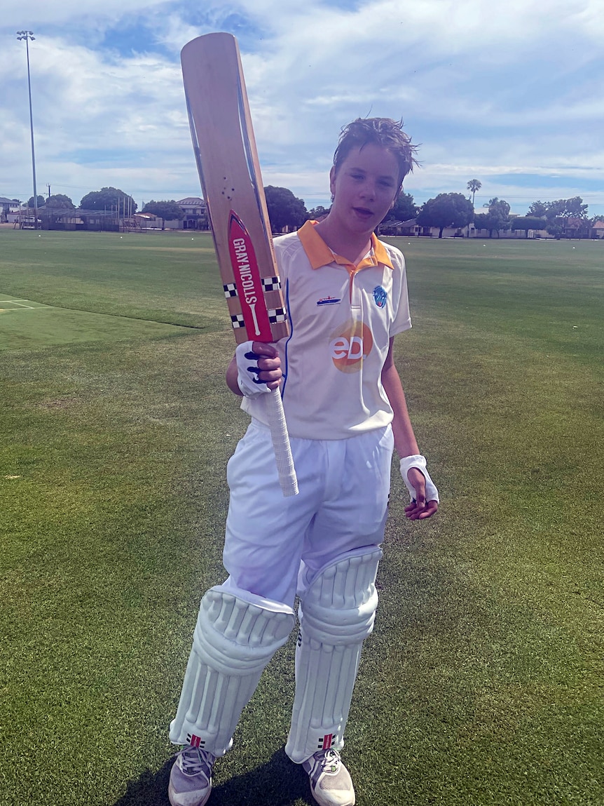 A young cricketer raises his bat as he walks off the ground.