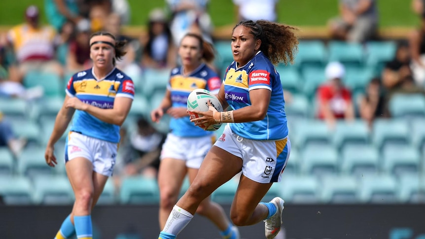 Three women play rugby in front of a crowd