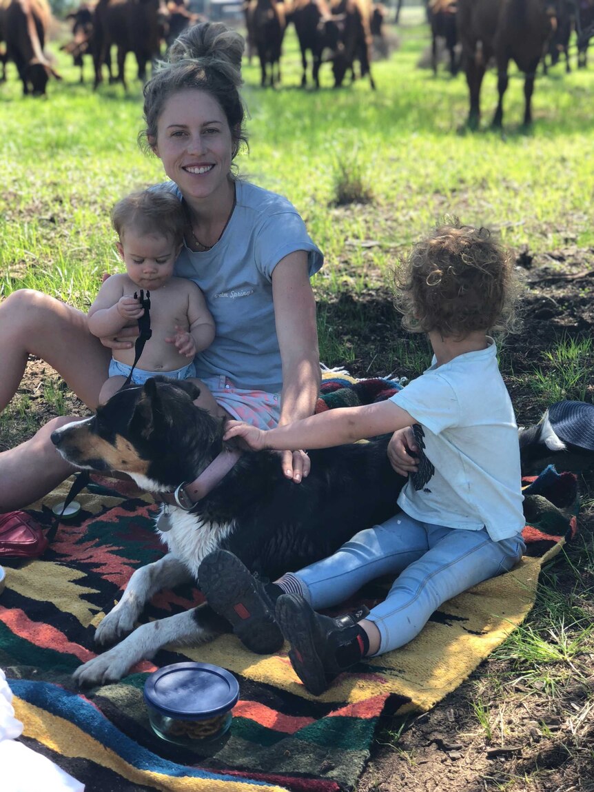 Medium shot of a woman sitting with children and a dog on a picnic rug in a paddock.