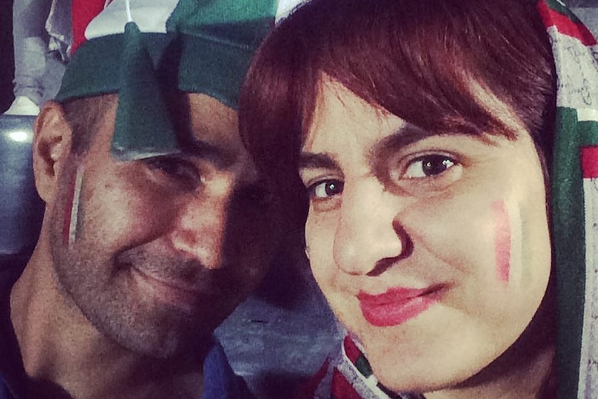 A woman in a red white and green heed scarf and facepaint smiled sitting next to a man in a jester's hat of the same colours.