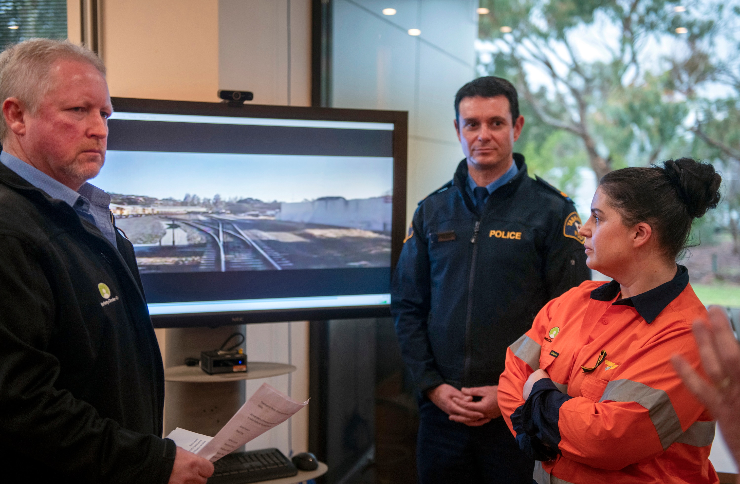 Two men and one woman in a fluro orange vest gather around a video screen depicting railroad tracks.