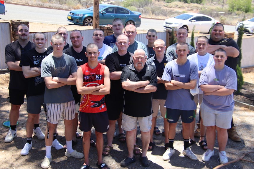 A group of young men and one older man with shaved heads standing in a cluster with their arms crossed