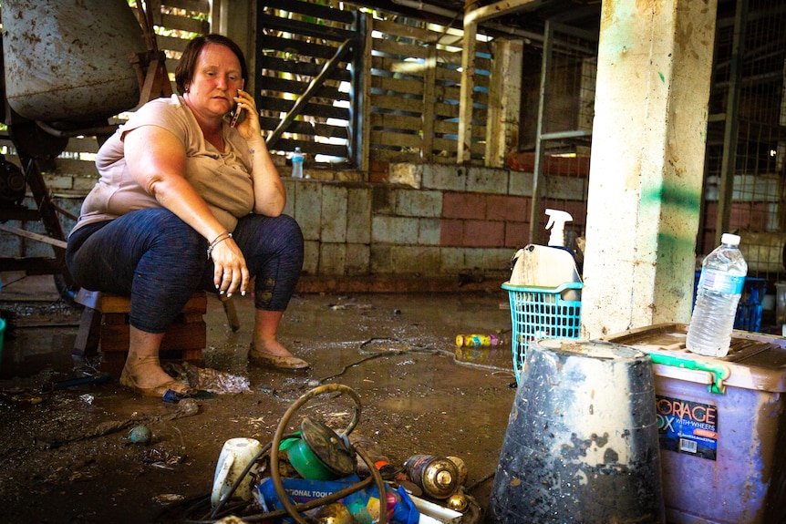 Kirsty MacGregor talks on her phone while sitting on a crate, on a muddy floor, surrounded by objectes caked in mud.