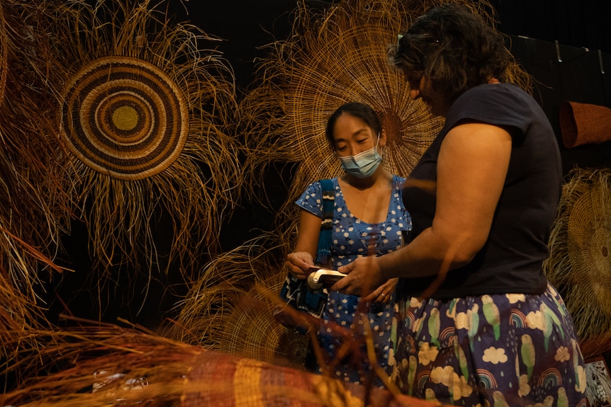 A woman scans her card on a card reader held by another woman in front of Pandanus mats hanging on a wall.